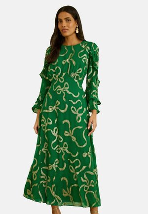 Frau trägt ein langes grünes Kleid mit goldbandähnlichen Mustern und gerafften langen Ärmeln, stehend vor einem einfarbigen Hintergrund.