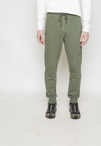 Pantalon de survêtement vert olive avec taille élastique et cordon de serrage, comprenant des poches latérales et des poignets fuselés. Associé à des chaussures de sport noires.