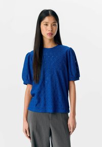 Blauwe kant blouse met korte pofmouwen, voorzien van ingewikkelde geometrische patronen en een ronde halslijn, gecombineerd met grijze wijde broek.