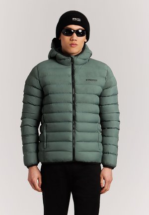 Hombre con chaqueta acolchada verde, gorro negro con logo, gafas de sol negras y pantalones negros de pie frente a un fondo claro y liso.
