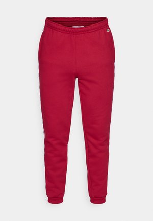 Red sweatpants made of soft fabric, featuring an elastic waistband and tapered cuffs. Side stripes display text in contrasting colors.