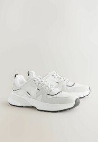 Baskets blanches en matériaux mesh et daim, avec des accents noirs, des lacets texturés, et une semelle blanche épaisse avec un embout gris.