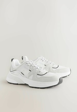 WeißeSportschuhe aus Netz und Wildledermaterialien, mit schwarzen Akzenten, strukturierten Schnürsenkeln und einer dicken weißen Sohle mit einer grauen Zehenkappe.
