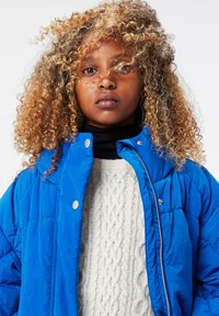 Blå puffjacka med dragkedja och tryckknappar, bärs över en krämfärgad stickad tröja med flätmönster. Lockigt, blont hår ramar delvis in ansiktet.