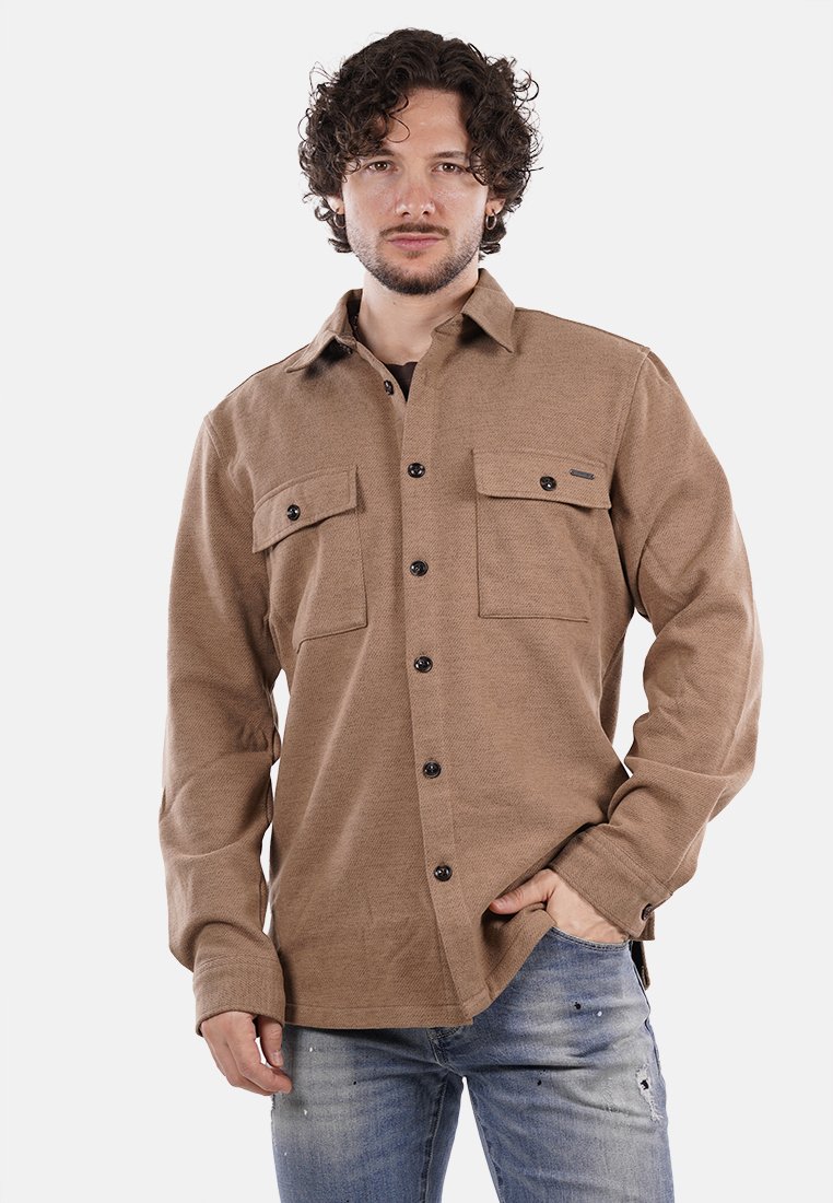 Camicia marrone con bottoni, realizzata in tessuto testurizzato, con due tasche sul petto, bottoni neri e colletto strutturato. Indossata con jeans blu.