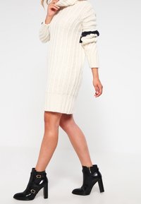 Knit cream sweater dress with a high collar, ribbed texture, and black stripe detail on sleeves. Paired with black ankle boots.