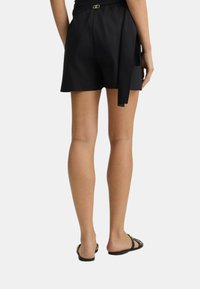 Pantaloni neri corti con dettagli in vita legati e passanti per cintura. Il tessuto appare liscio; design minimalista con un taglio dritto. Indossati con sandali neri.