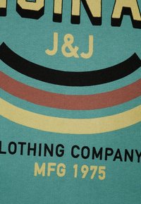 Tessuto turchese con trama lavorata a maglia, caratterizzato da strisce audaci nere, bordeaux, gialle e turchesi, e testo: "J&J Clothing Company MFG 1975."