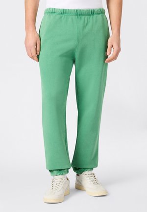 Osoba ubrana w zielone spodnie dresowe z elastycznym pasem i kremowo-białe trampki, stojąca na tle jednolitego jasnego tła.