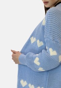 Maglione lavorato a maglia azzurro chiaro con motivi di cuori bianchi, polsini a costine e una vestibilità comoda. La texture sembra morbida e di peso medio.