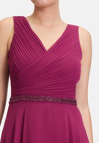 Fukszia ruha plisszírozott mellel, mély V-nyakkal és gyöngyös díszítéssel a derékon. Simább anyagból készült, bővülő szoknyával és ujjatlan kivitelben.