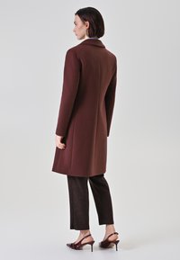 Cappotto di lana marrone con una vestibilità su misura, maniche lunghe e colletto a revers, abbinato a pantaloni scuri a fantasia e scarpe con tacco bordeaux.
