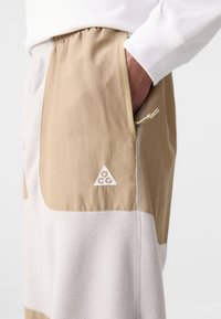 Beige and gray color-blocked joggers made of soft fleece with an elastic waistband and side zip pocket, featuring a small white logo.