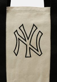 Sac tote avec un corps crème, des poignées noires et un logo "NY" noir brodé. Fabriqué en toile durable avec une texture lisse.