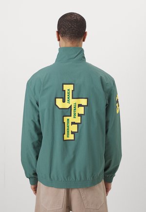 Grüne leichte Jacke mit hohem Kragen und elastischen Bündchen, mit gelb-schwarzen Grafiken auf dem Rücken, die "JAMAICA FOOTBALL FEDERATION" lesen.