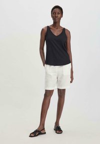 Top de tirantes negro con escote en V y una textura suave, combinado con pantalones cortos de lino blancos que tienen dos bolsillos frontales. Lleva sandalias negras con nudo.