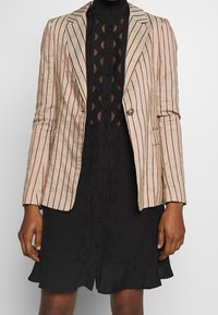 Blazer a righe beige e rosso, con design aderente, chiusura con un bottone e spalle strutturate; abbinato a un vestito nero strutturato.