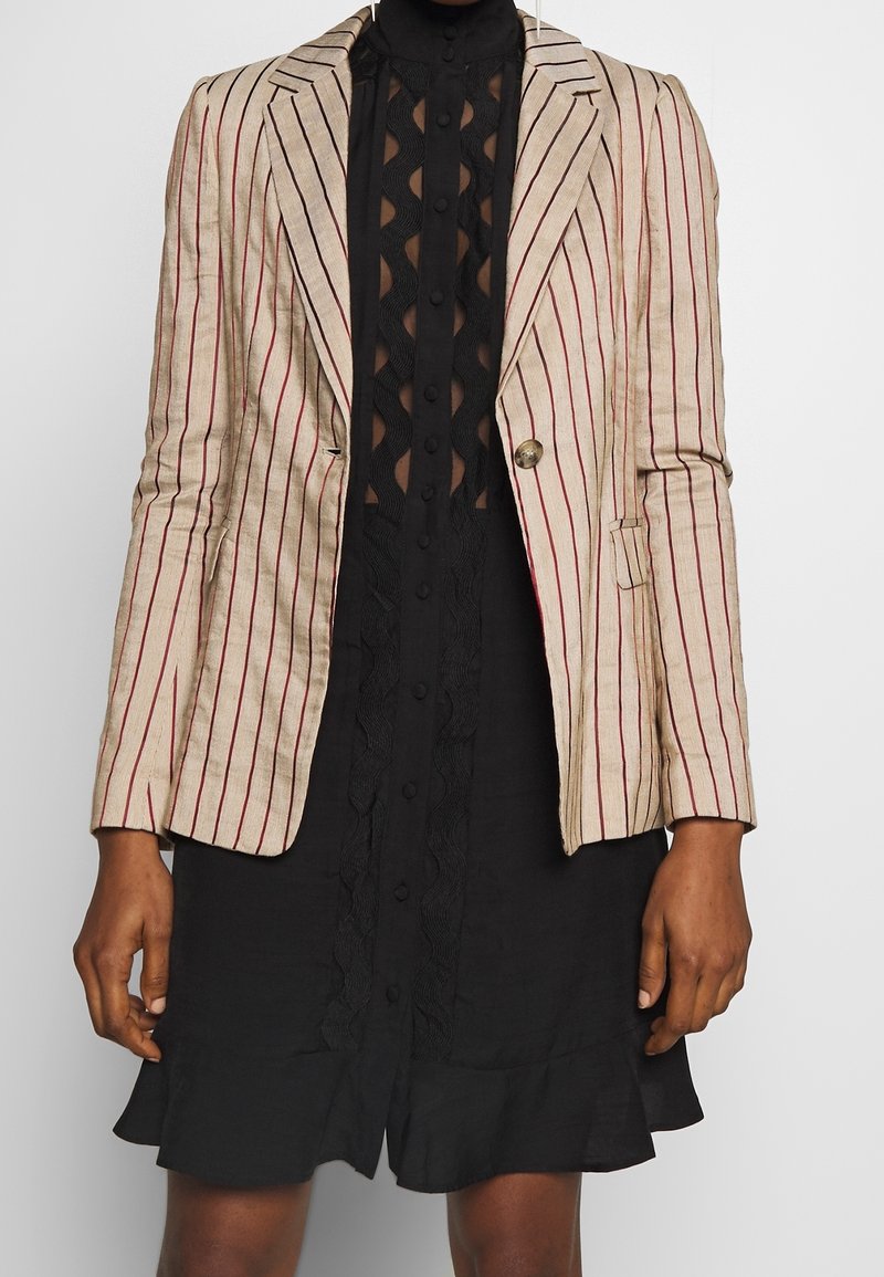 Blazer a righe beige e rosso, con design aderente, chiusura con un bottone e spalle strutturate; abbinato a un vestito nero strutturato.