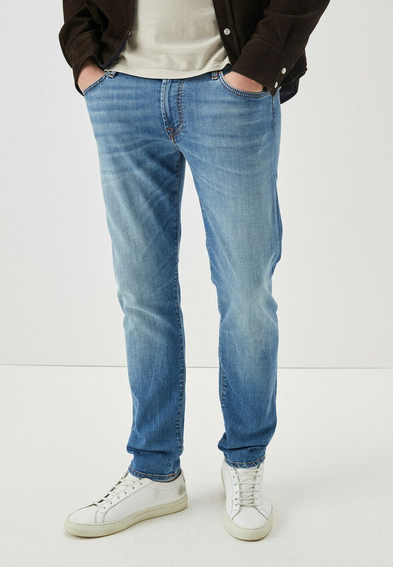 Jeans in denim blu chiaro con vestibilità slim, leggere sbiaditure, tasche anteriori e chiusura con bottone, abbinati a sneaker bianche.