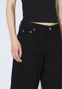Pantalones de mezclilla negros con corte recto, que cuentan con herrajes de cobre, cinco bolsillos y un cierre de botón en la cintura.