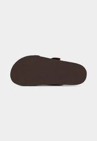 Semelle d'une sandale brun foncé avec un dessous plat et texturé et une seule sangle visible au-dessus, sur un fond gris clair.