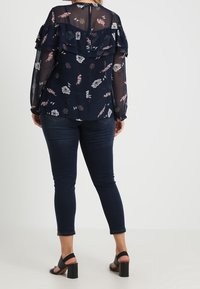 Marinebluse mit floralem Druck, gestuftem Rüschendesign, langen durchsichtigen Ärmeln und einem Schlüssellochdetail im Rücken, kombiniert mit dunklen Jeans und schwarzen Blockabsätzen.