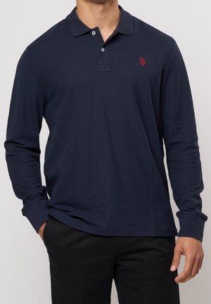 Mann trägt ein marineblaues Langarm-Polohemd mit gesticktem roten Logo und schwarze Hose, Hände entspannt an den Seiten, vor einfarbigem Hintergrund.