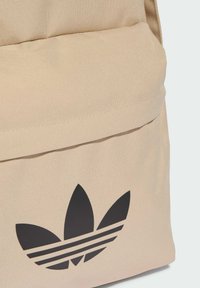 Beżowy plecak wykonany z teksturowanej tkaniny, z wyraźnym czarnym logo trójlistka Adidas oraz wzorem trzech pasków na przedniej kieszeni.