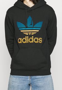 Osoba v černé mikině s kapucí a modro-oranžovým pruhovaným logem Adidas, spárovaná se světle modrými džíny, na jednobarevném pozadí.