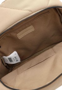 Beigā rokassomas iekšpuse, kur redzama kabata ar rāvējslēdzēju, auduma birka ar zīmolu un balta produkta etiķete galvenajā nodalījumā.