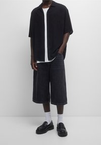 Camicia nera a maniche corte, t-shirt bianca sotto, pantaloni corti in denim scuro fino al ginocchio, mocassini neri con suola spessa, calzini bianchi.