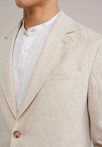 Beige Fischgrätenblazer mit einem einzelnen Holzknopf, ausgestattet mit einer Brusttasche und einem strukturierten weißen Hemd darunter.