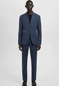 Mężczyzna stojący, ubrany w dopasowany ciemnoniebieski garnitur, czarne buty i ciemną podkoszulkę, na tle jednolitego, jasnoszarego tła.