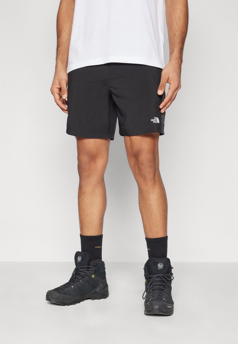 Sorte idrettsshorts med en avslappet passform, med en liten hvit logo på venstre bein, sammen med svarte tursko og mørke sokker.