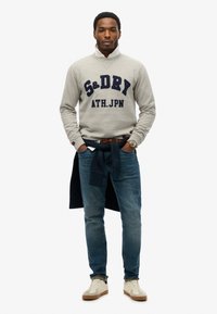 Sudadera gris con texto azul marino "S&DRY ATH.JPN," combinada con jeans azules y zapatillas de color claro. Este conjunto presenta un estilo casual y en capas.