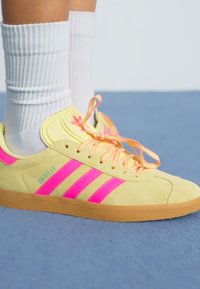 Dzelteni zamšaini sporta apavi ar rozā svītrām, oranžām šņorēm un teksturētu gumijas zoli. Iespēts logo un mīkstināta apkakle. Redzami baltie sporta zeķi.