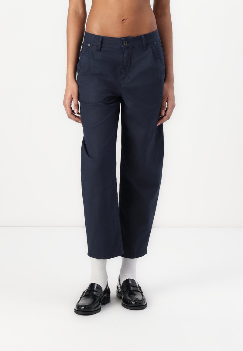 Navy katoen broek met een relaxte pasvorm, voorzien van voorzakken, een knoopsluiting en contrasterende zwarte instappers gedragen met witte sokken.