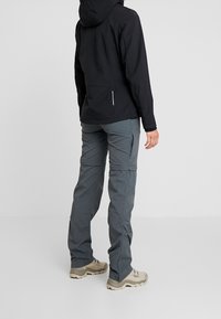 Veste noire imperméable avec capuche et poches zippées, associée à un pantalon cargo gris. Chaussures au design robuste.