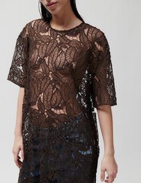 Γυναίκα που φοράει διάφανο καφέ δαντελωτό τοπ με μεγάλα φυλλώδη σχέδια πάνω από μπλε τζιν, στέκεται μπροστά σε απλό φόντο.