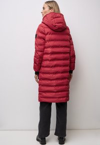 Donna con cappotto imbottito lungo rosso con cappuccio, in piedi di lato, abbinato a pantaloni neri e scarpe nere su sfondo semplice.