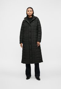 Long manteau matelassé noir avec un design en losanges, des boutons à l'avant et une capuche. Le tissu semble lisse et isolé, adapté aux temps froids.