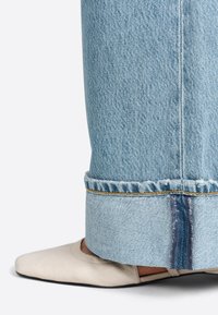 I jeans in denim azzurro chiaro presentano un orlo sfilacciato con una striscia blu a contrasto e un accento di cucitura gialla. Un mocassino beige con punta è parzialmente visibile.