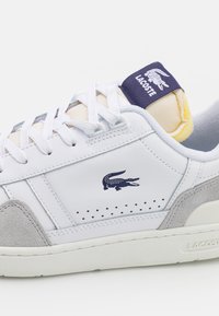 Baskets en cuir blanc avec des accents en daim gris, dotées d'un design perforé, de lacets, et d'une languette bleu marine arborant le label "LACOSTE".