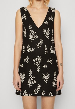 Czarna sukienka bez rękawów z głębokim dekoltem w kształcie litery V i białym kwiatowym haftem, noszona przez osobę z małym tatuażem serca na lewej przedramieniu.