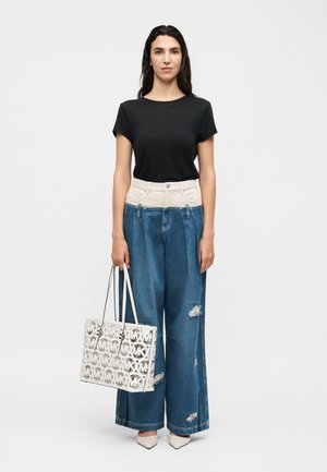 Mujer de pie frente a fondo blanco, viste camiseta negra, vaqueros anchos de talle alto con rotos, zapatos blancos puntiagudos, sosteniendo un bolso tote blanco con estampado.