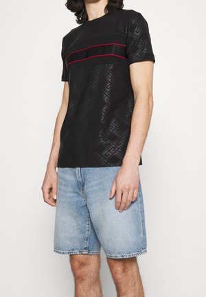 Mężczyzna ubrany w czarną koszulę z krótkim rękawem w wzory i czerwonymi paskami na wysokości klatki piersiowej oraz jasnoniebieskie krótkie dżinsy.