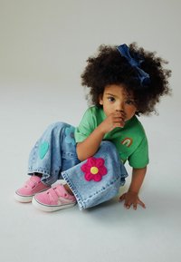 T-shirt vert à manches courtes avec détail arc-en-ciel, pantalon large en denim bleu orné d'appliqués colorés en feutre en forme de fleurs et de cœurs, et baskets roses.