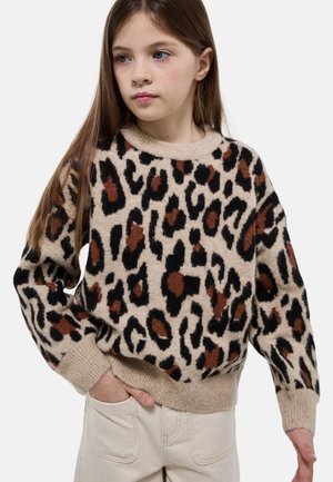 Jeune fille aux longs cheveux bruns portant un pull imprimé léopard beige, marron et noir, ainsi qu'un pantalon clair, la main dans la poche.