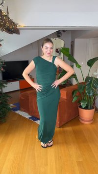 Πράσινο, εφαρμοστό φόρεμα με ασύμμετρο ντεκολτέ και ρυτιδωτή υφή. Η μοντέλο φοράει σαγιονάρες και ποζάρει σε ένα σαλόνι με φυτά και έπιπλα.