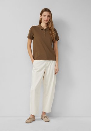 Femme debout contre un mur gris portant un polo marron à manches courtes, un pantalon blanc à jambes larges et des chaussures beige à lacets.